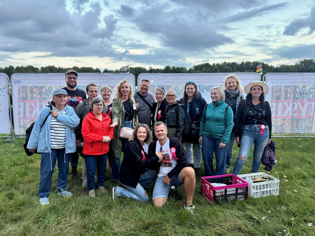 Fifteen people pose as a group in front of a fence with Pol’and’Rock banners. Two people kneel in front. The scene includes grassy ground and a cloudy sky.